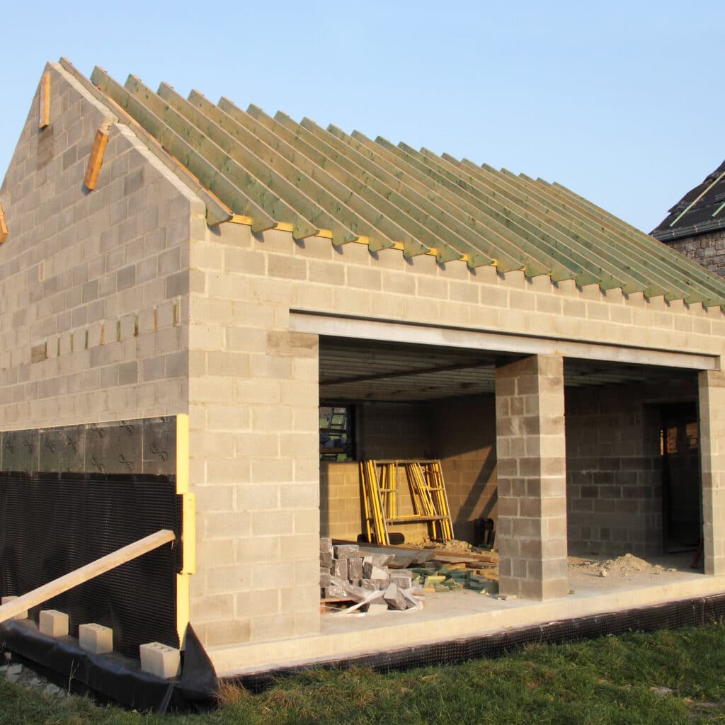 Bâtiment en construction : garage en parpaings avec charpente bois posée. Isolation RECTICEL visible en soubassement. Matériaux de chantier.
