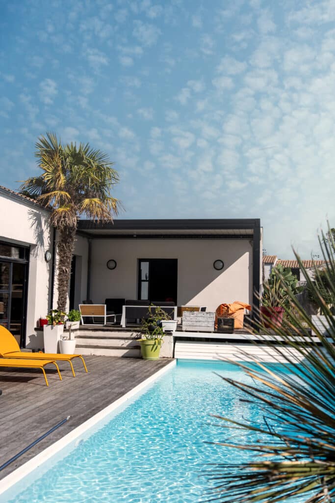 Piscine et terrasse en bois: Détente estivale à la maison Maison moderne avec piscine bleue, terrasse en bois, palmier et chaise longue jaune sous un ciel clair. Ambiance estivale et relaxante.