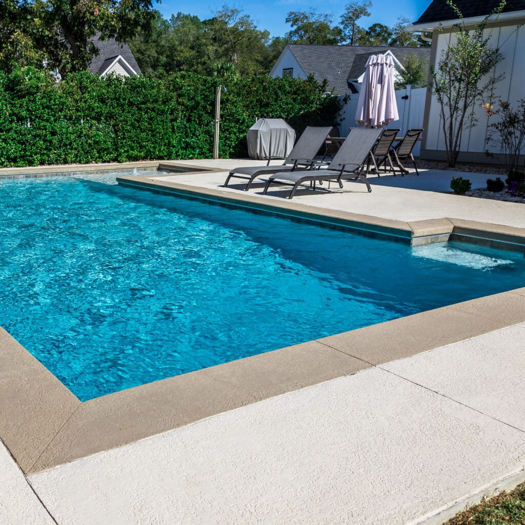 Piscine moderne avec spa et terrasse en béton Piscine rectangulaire bleue avec spa intégré, entourée d'une terrasse en béton clair. Chaises longues et parasol fermé dans un jardin verdoyant avec une maison visible en arrière-plan.
