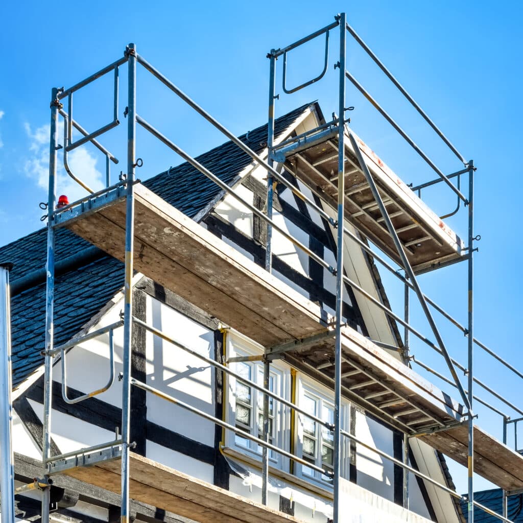 Rénovation maison à colombages : échafaudage et ciel bleu Échafaudage métallique et planches devant une maison à colombages blanche et noire en rénovation. Toit en tuiles foncées, ciel bleu lumineux.