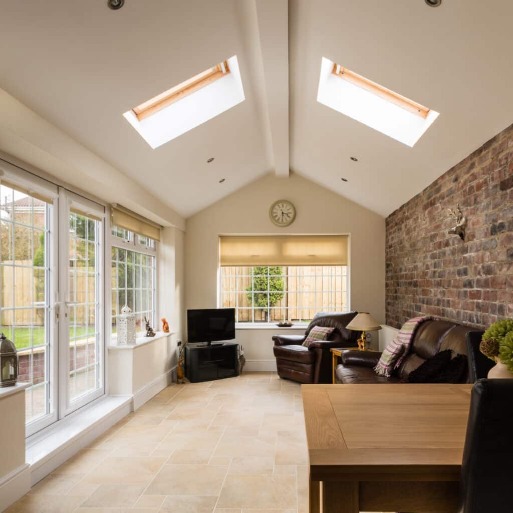 Véranda lumineuse avec puits de lumière, salon confortable en cuir, mur en briques apparentes et vue sur un jardin clôturé.